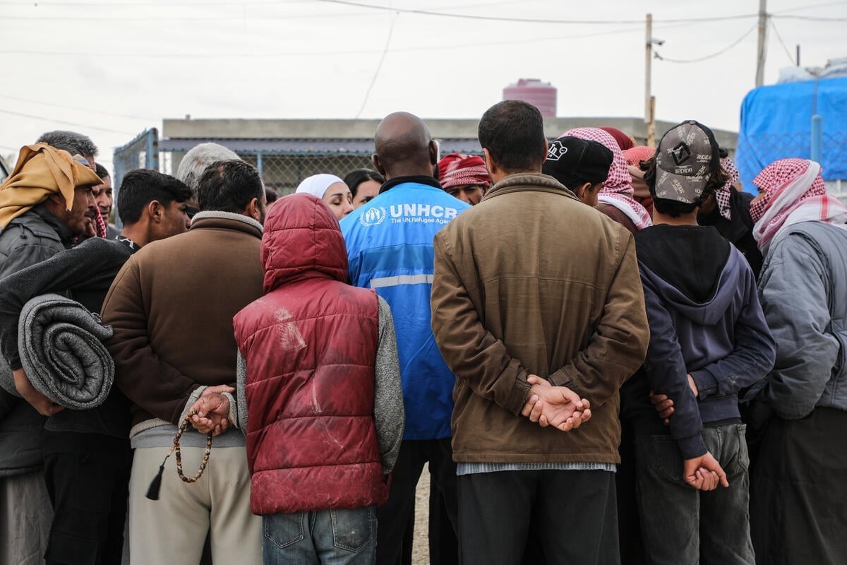 Rear view of a man wearing a blue UNHCR vest surrounded by a small group of people outside in a camp.