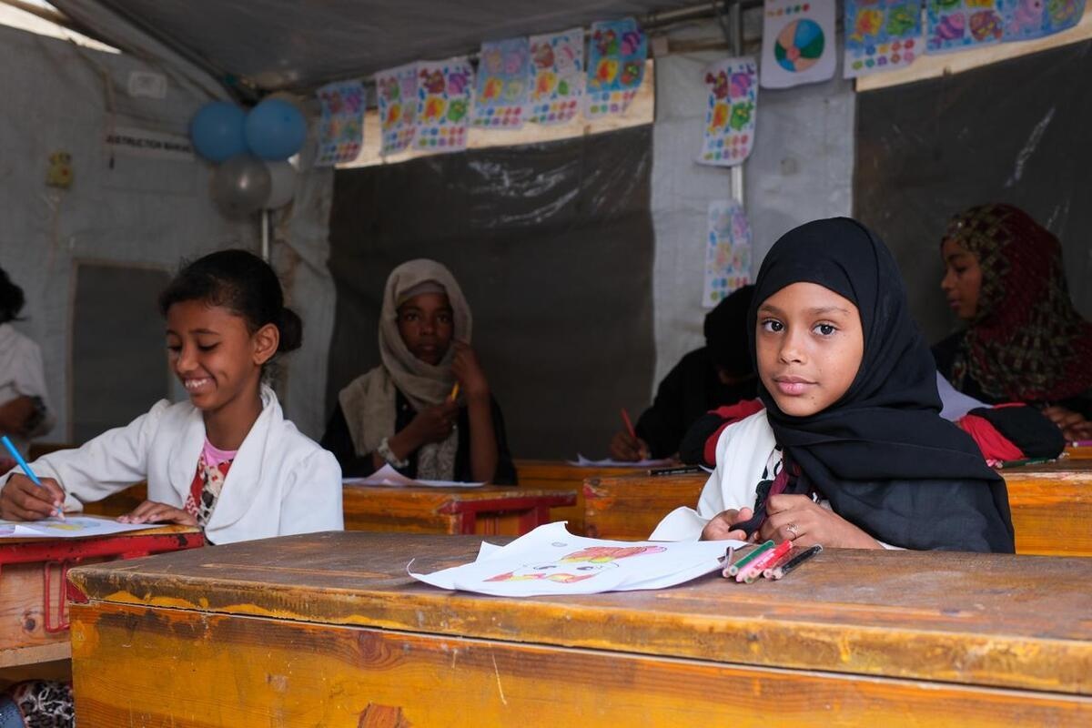 Girls sit at wooden desks with paper and coloured pencils in classroom inside a tent.