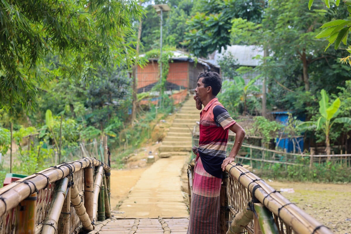 A refugee with a disability stands on a bridge made from bamboo in a camp.