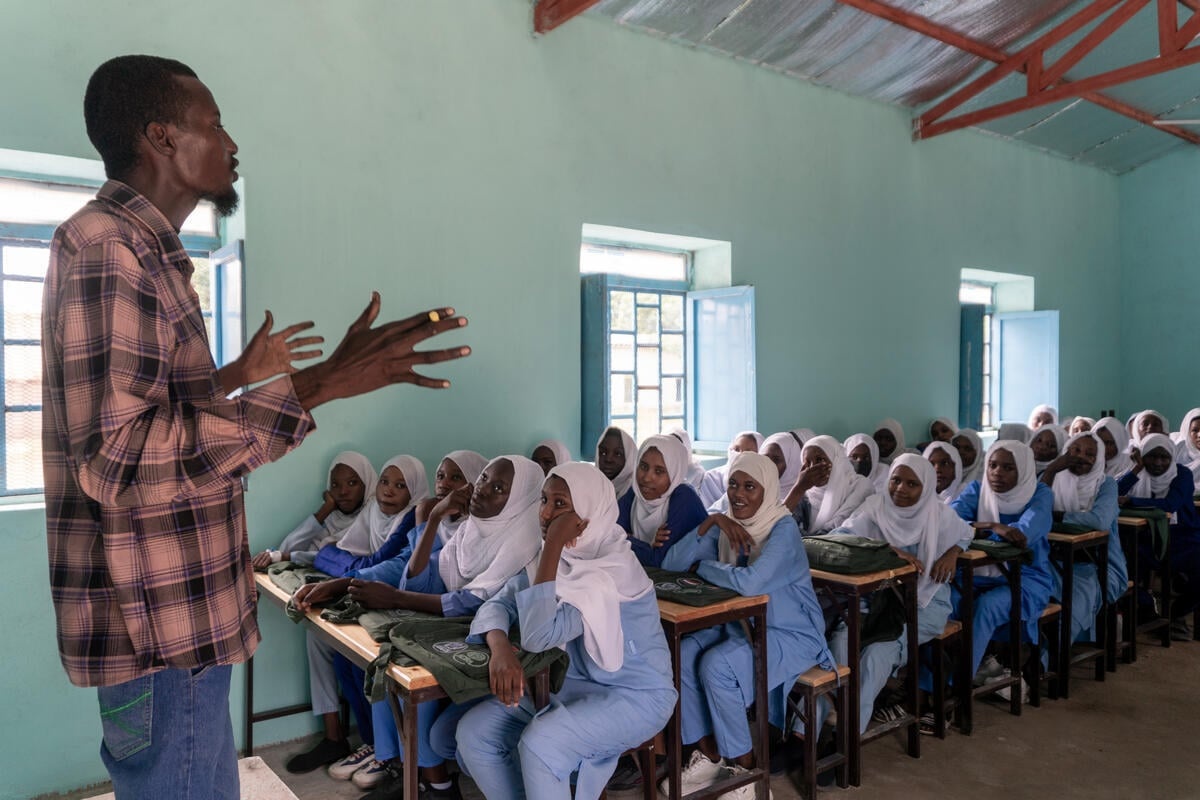 A teacher wearing a checked shirt and jeans stands at the front of a classroom addressing schoolgirls seated behind rows of desks