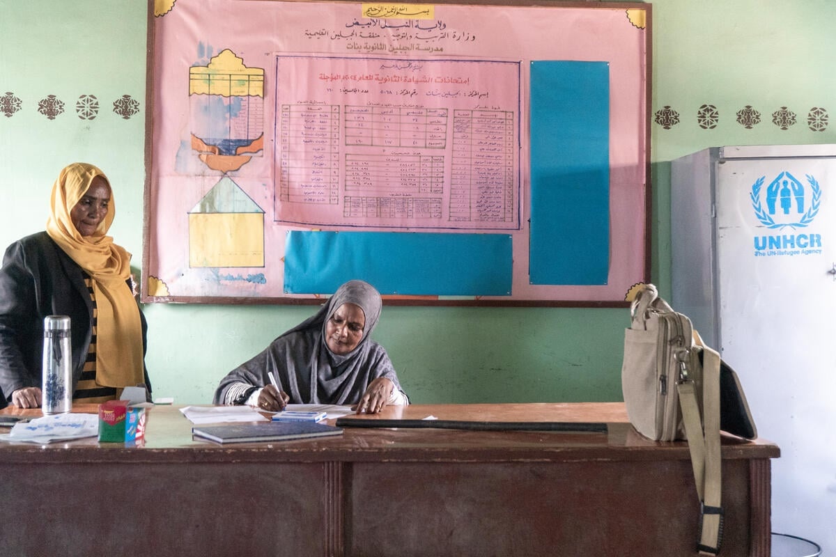 A woman sits behind a teacher's desk writing at the front of a classroom as another woman stands on her right