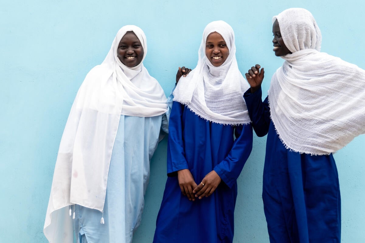 Three schoolgirls wearing blue dresses and white headscarves smile while standing in front of a turquoise wall