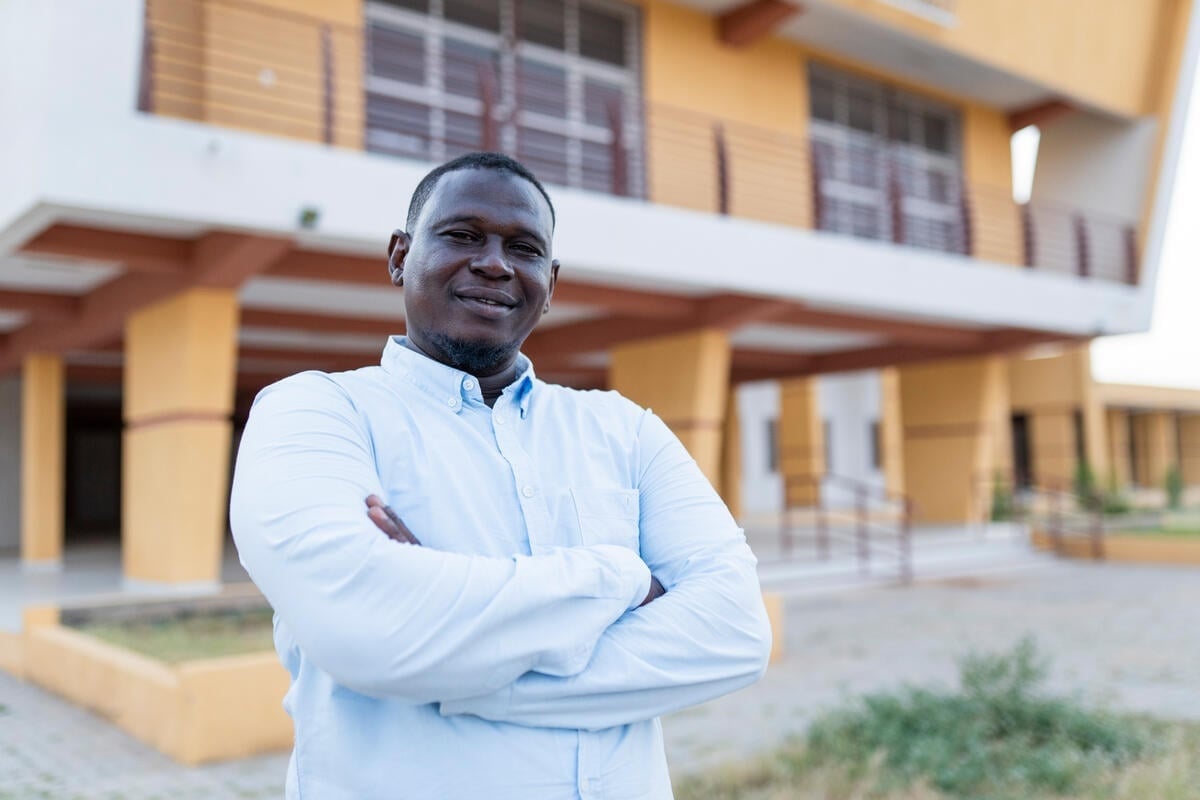 A smiling man wearing a pale button down shirt stands outside with his arms crossed in front of a yellow-painted building