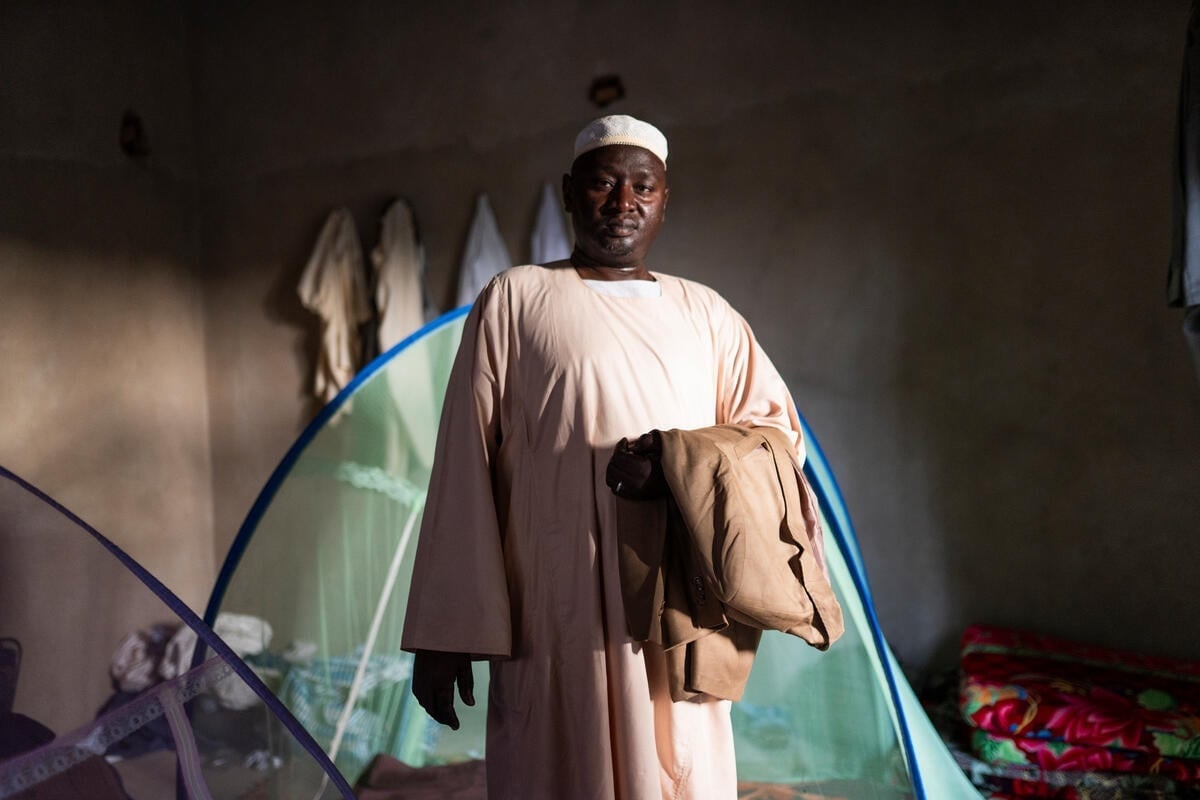 A man stands holding a folded suit over his left forearm in a shaded room with mosquito net tents in the background