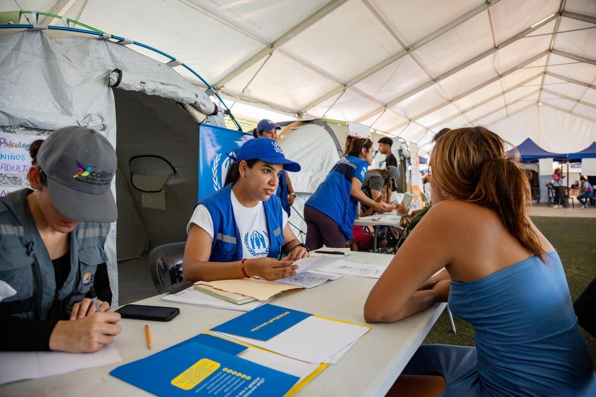 Humanitarian workers sitting behind tables meet with people inside a large tent