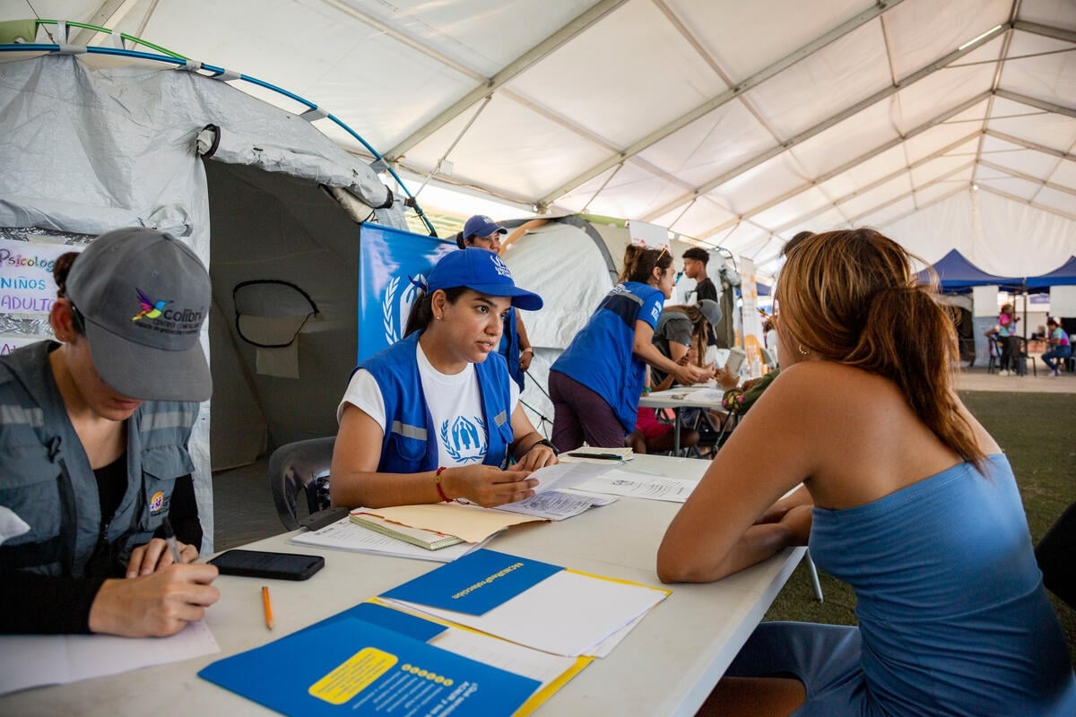 Humanitarian workers sitting behind tables meet with people inside a large tent