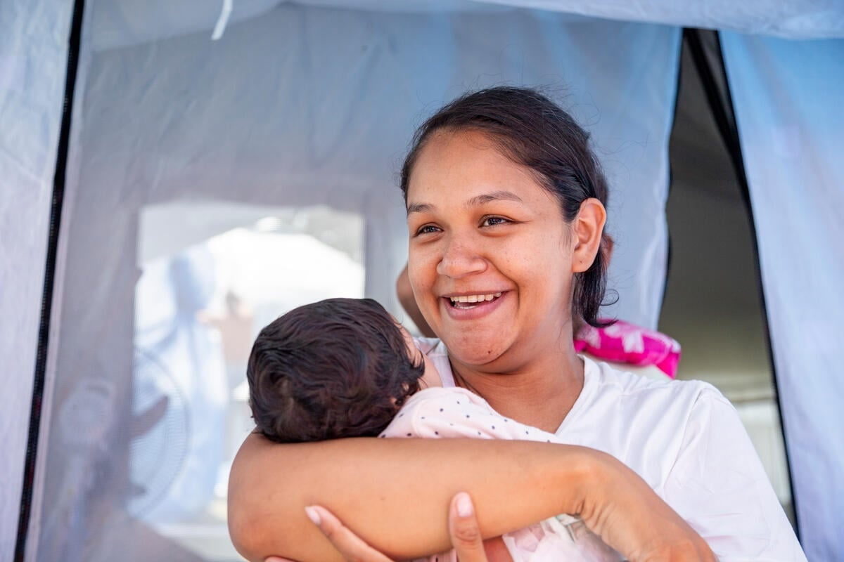 A smiling woman cradles a baby in her arms in front of a tent opening.