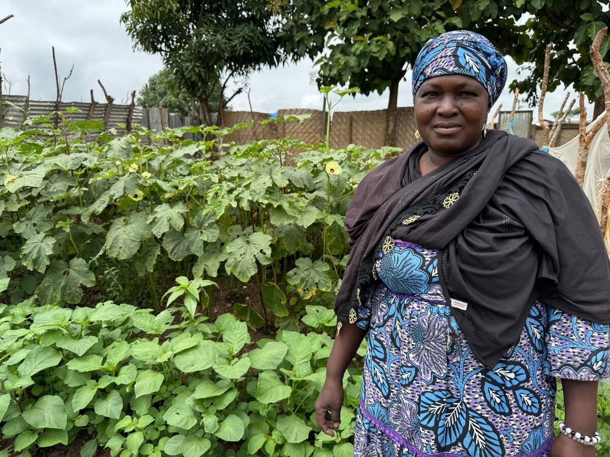 A woman wearing a floral robe and matching headscarf stands in front of a garden filled with plants