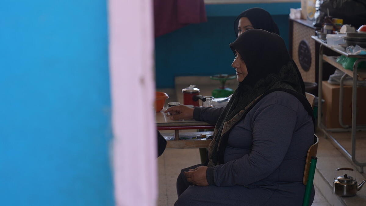 Two women wearing headscarves sit on school chairs in a classroom with a kettle and gas cooker visible on the floor 