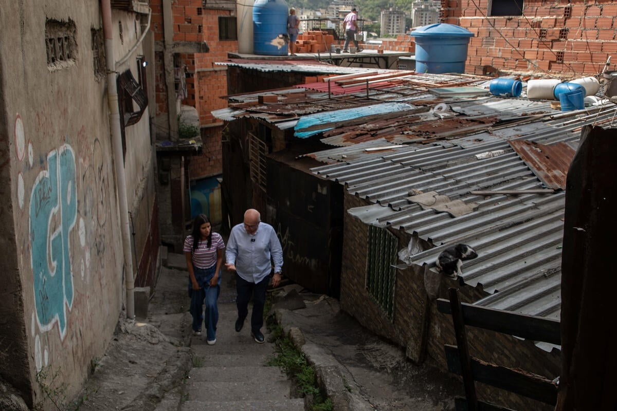 A man and a woman walk side by side up the staircase of a narrow street beside tin-roofed buildings 