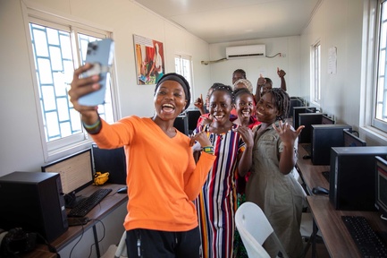 A woman in a bright orange shirt takes a selfie with a group of young women in a computer lab.