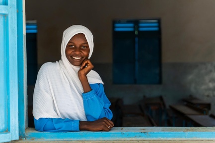 A young girl smiles at the camera, looking through an open window.