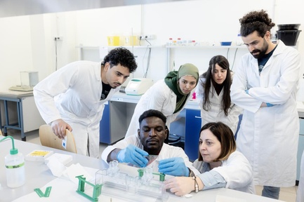 A group of people wearing lab coats gather round as a student and professor carry out an experiement in a laboratory 