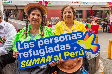 Two smiling people hold up a sign which reads 'Las personas refugiadas suman al Perú'. Behind them a festival is underway.
