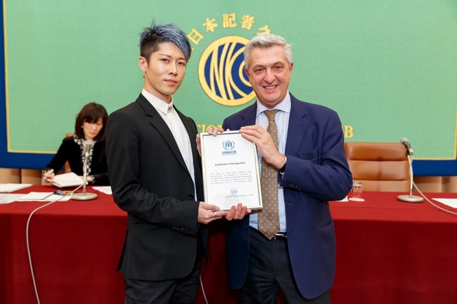 MIYAVI stands with Filippo Grandi and a certificate they are holding between them.