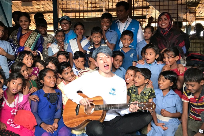 A man pictured playing a guitar, with Rohingya refugee children gathered closely behind him. Some adults and UNHCR staff stand at the back.