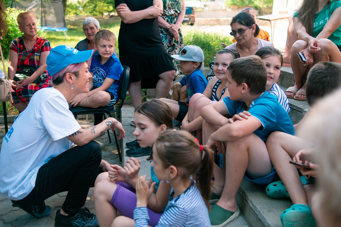 MIYAVI is crouched on the ground, smiling, a group of Ukrainian refugees around him.