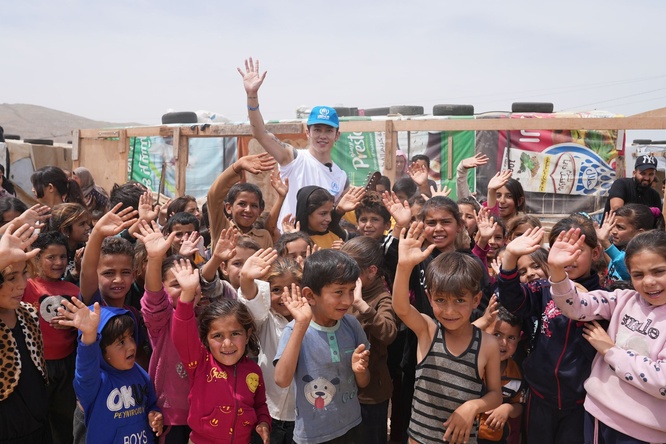 MIYAVI stands with his guitar, waving, behind a large group of refugee children, also waving.