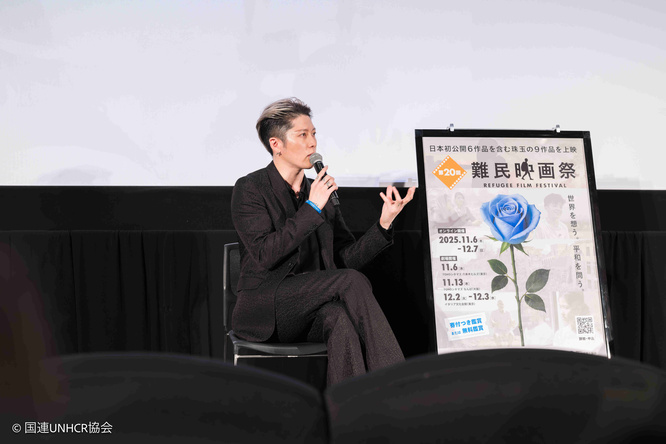 MIYAVI speaks on stage while seated beside a poster for the 20th Refugee Film Festival, Japan.