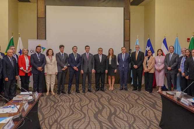 Un grupo de 18 hombres y 11 mujeres con vestimenta formal, de pie en una sala de juntas, detrás de ellos se encuentran 13 banderas coloridas de diferentes países
