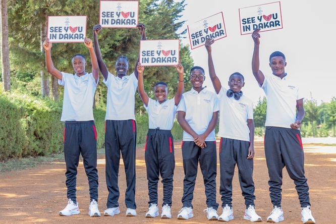 Six smiling young athletes wearing training shoes and clothes hold up signs reading 'see you in Dakar' on a wide dusty road 