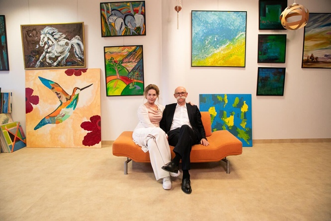 A woman and a man pose on an orange sofa, surrounded by art