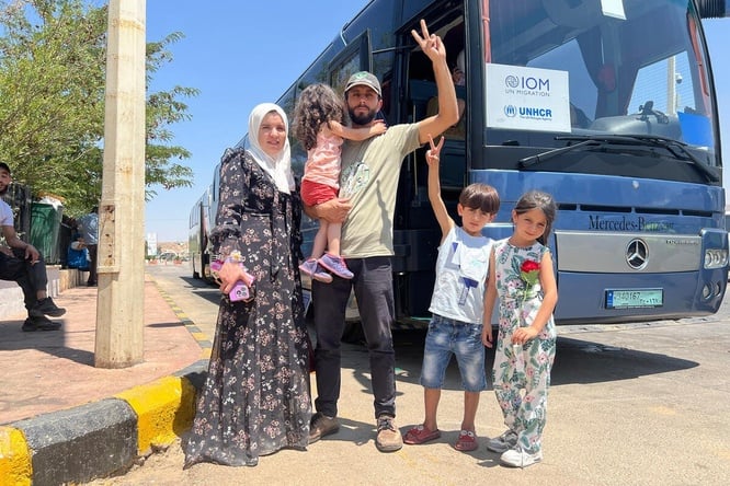 A couple and their three children stand on the roadside, smiling and waving in front of a bus with a UNHCR and IOM sign in the window. 