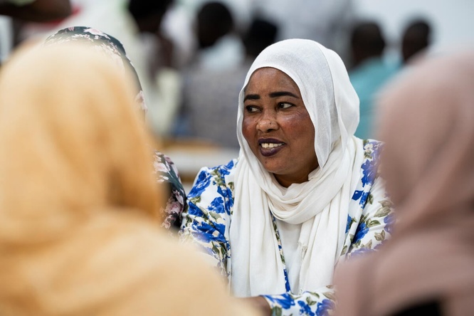 A smiling woman in a white headscarf and blue floral dress sits at a table with other women