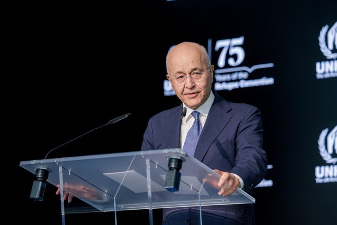 A man in a suit delivers a speech standing at a clear lectern in front of a dark background.