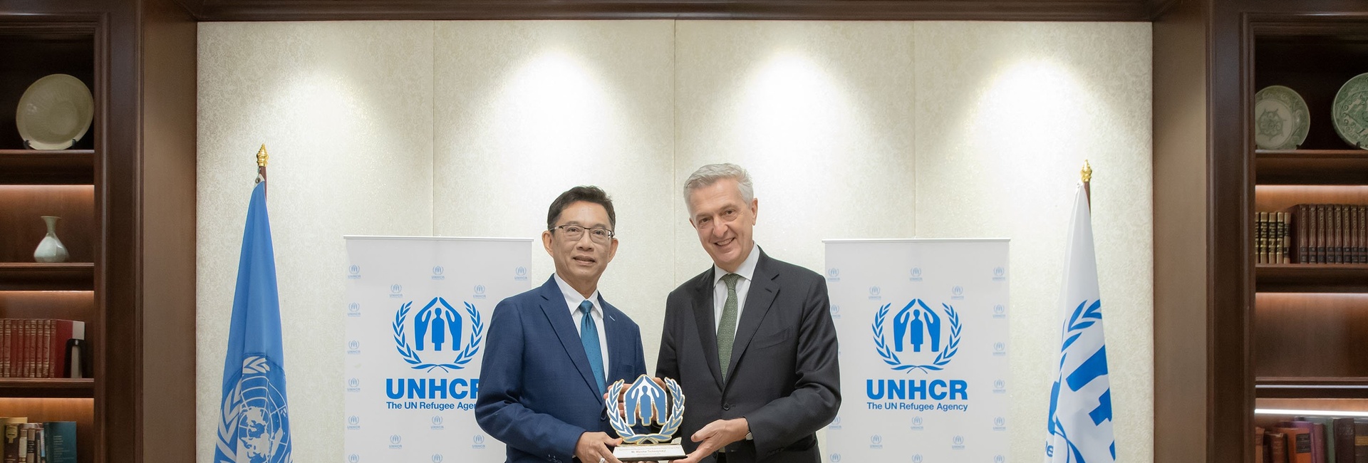 Mr. Wanchai Tachavejnukul and Filippo Grandi smiling and holding an award together, standing in front of UNHCR banners and flags