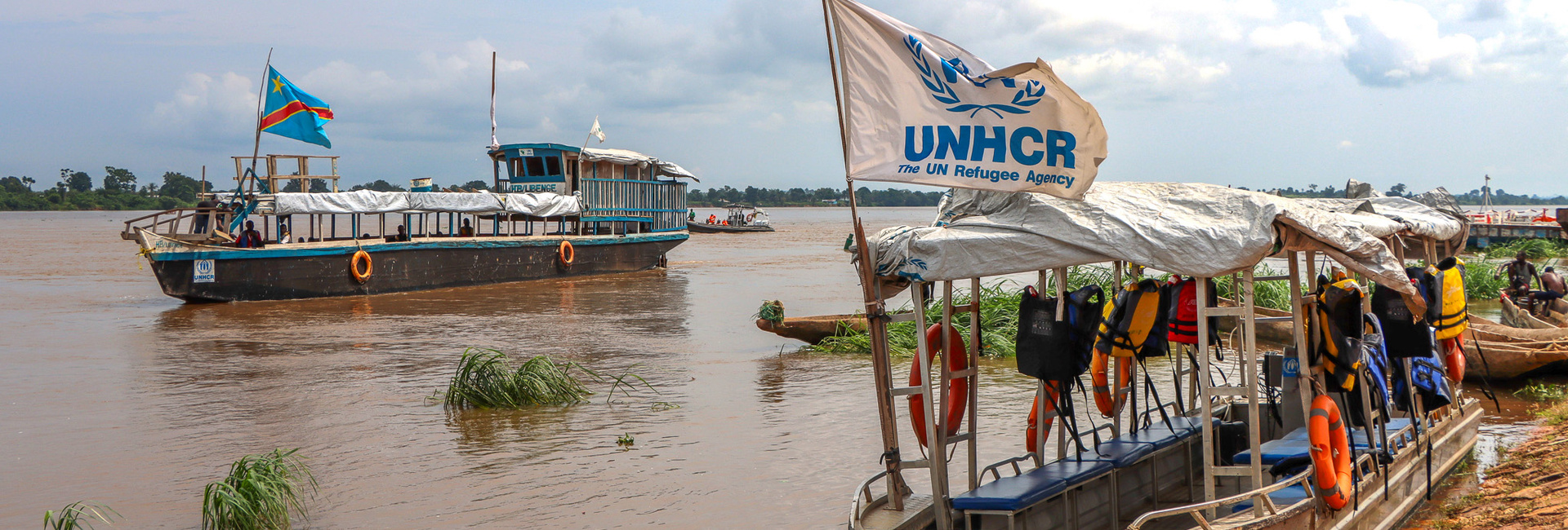 Wooden boats float on a wide, muddy river. One bears a UNHCR flag.