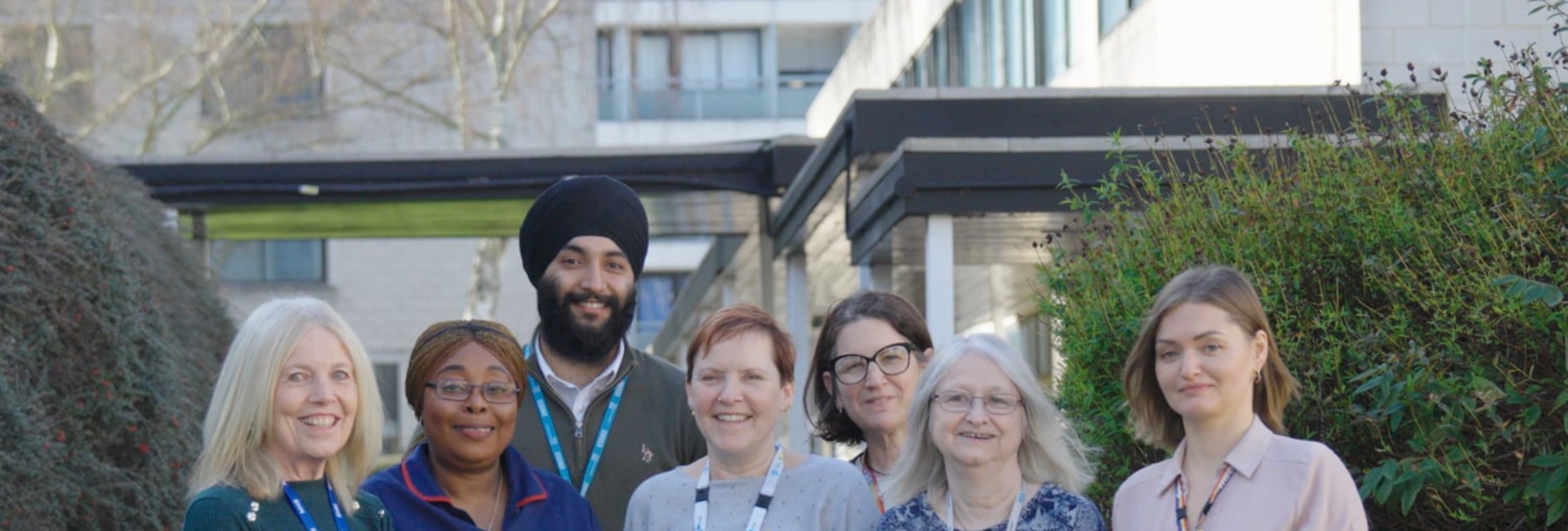 A group of six women and one man, most wearing hospital lanyards and one in a nurse's uniform, stand outside a building flanked by bushes