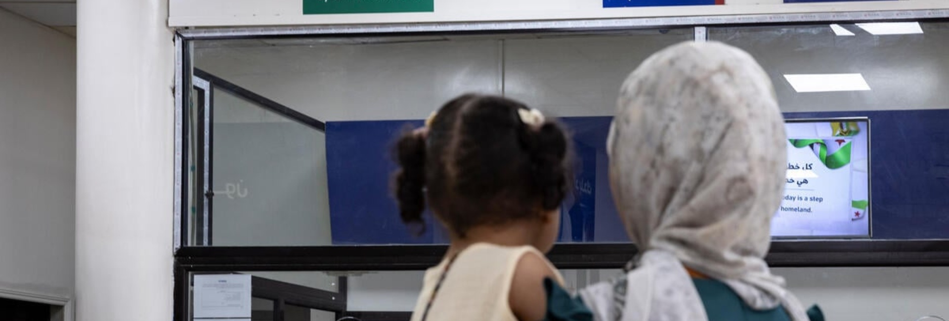 A woman carrying her daughter stands in front of a reception counter with a sign reading 'Syrian returnees' in Arabic and English
