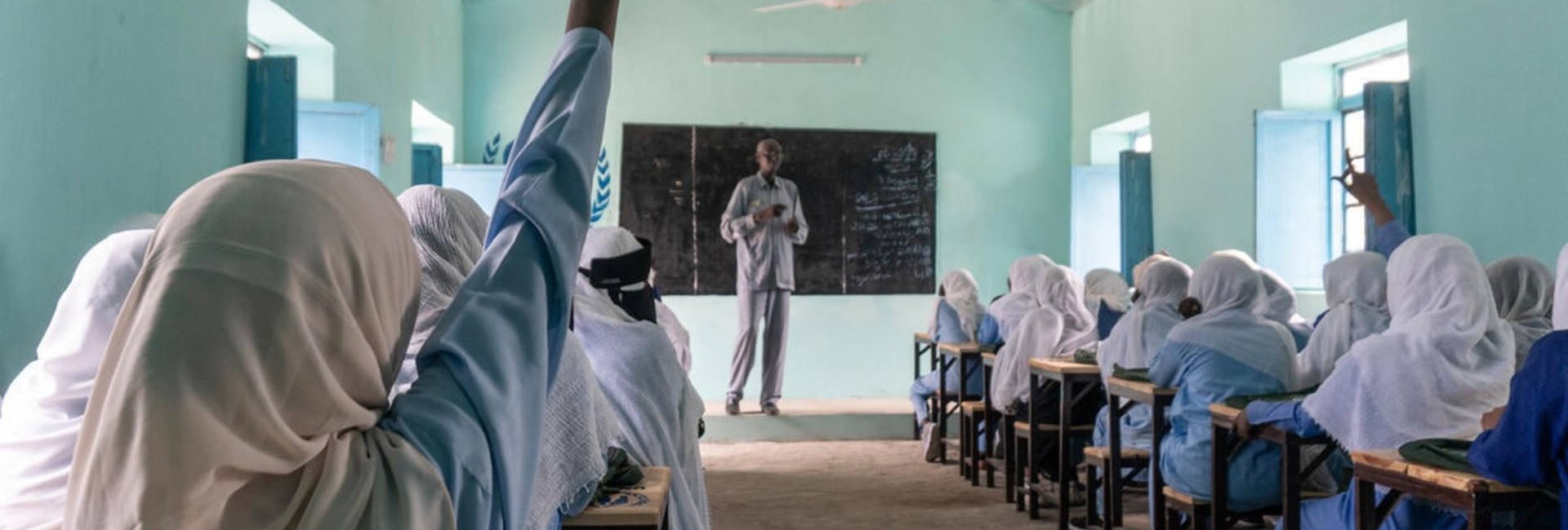 A rear view of a pupil raising her hand at the back of a classroom filled with schoolgirls in uniform sitting at rows of desks 