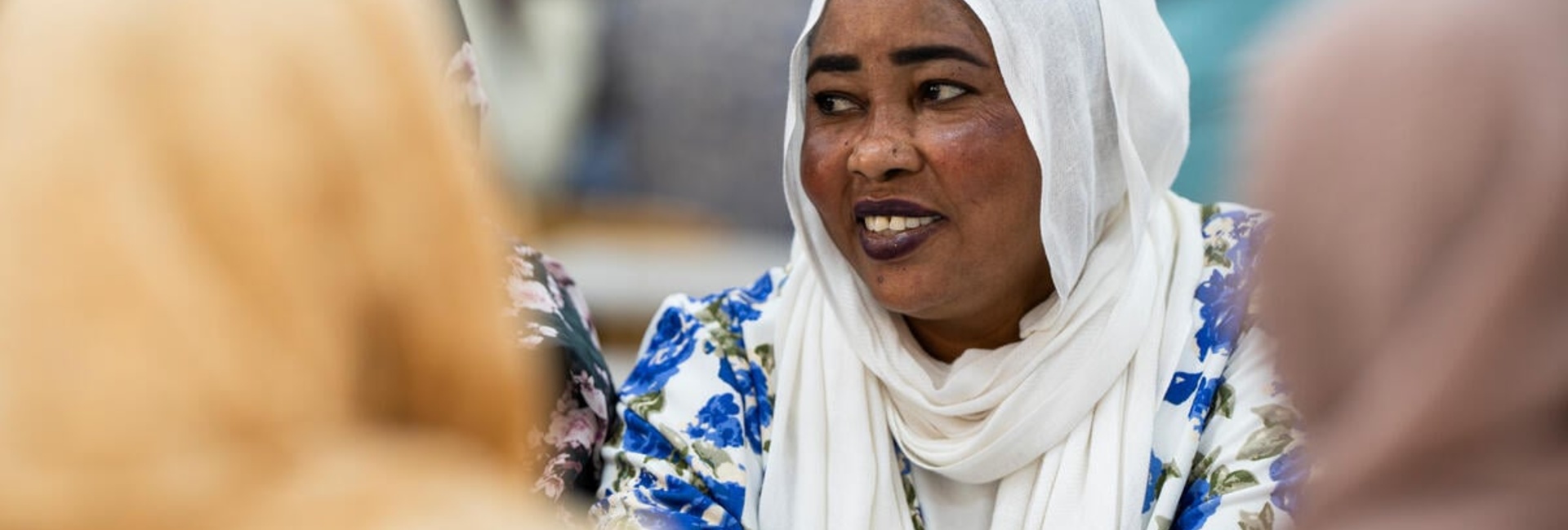 A smiling woman in a white headscarf and blue floral dress sits at a table with other women