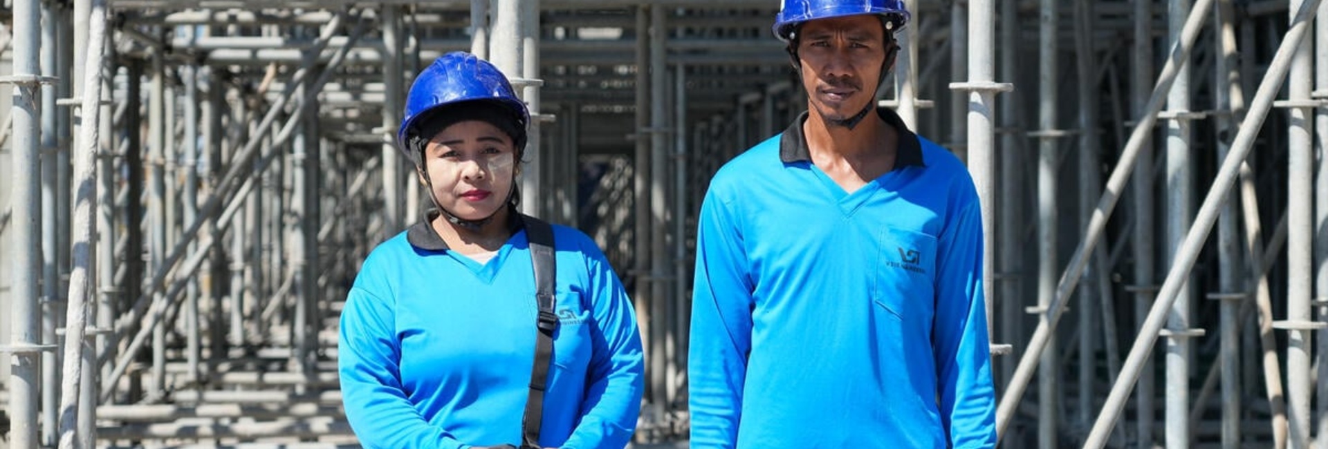 A woman and a man wearing matching blue shirts and hard hats stand outside in front of scaffolding