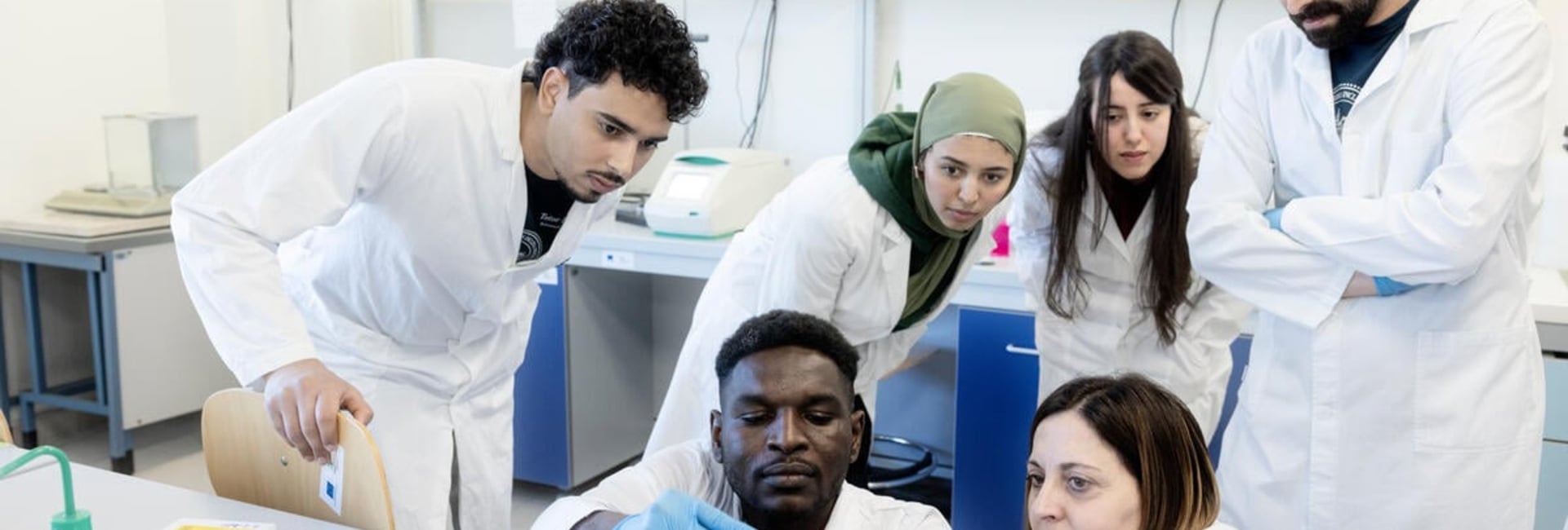 A group of people wearing lab coats gather round as a student and professor carry out an experiement in a laboratory 