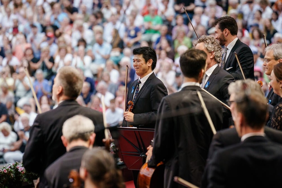 Musicians part of an orchestra stand up holding their instruments, in front of a crowd.