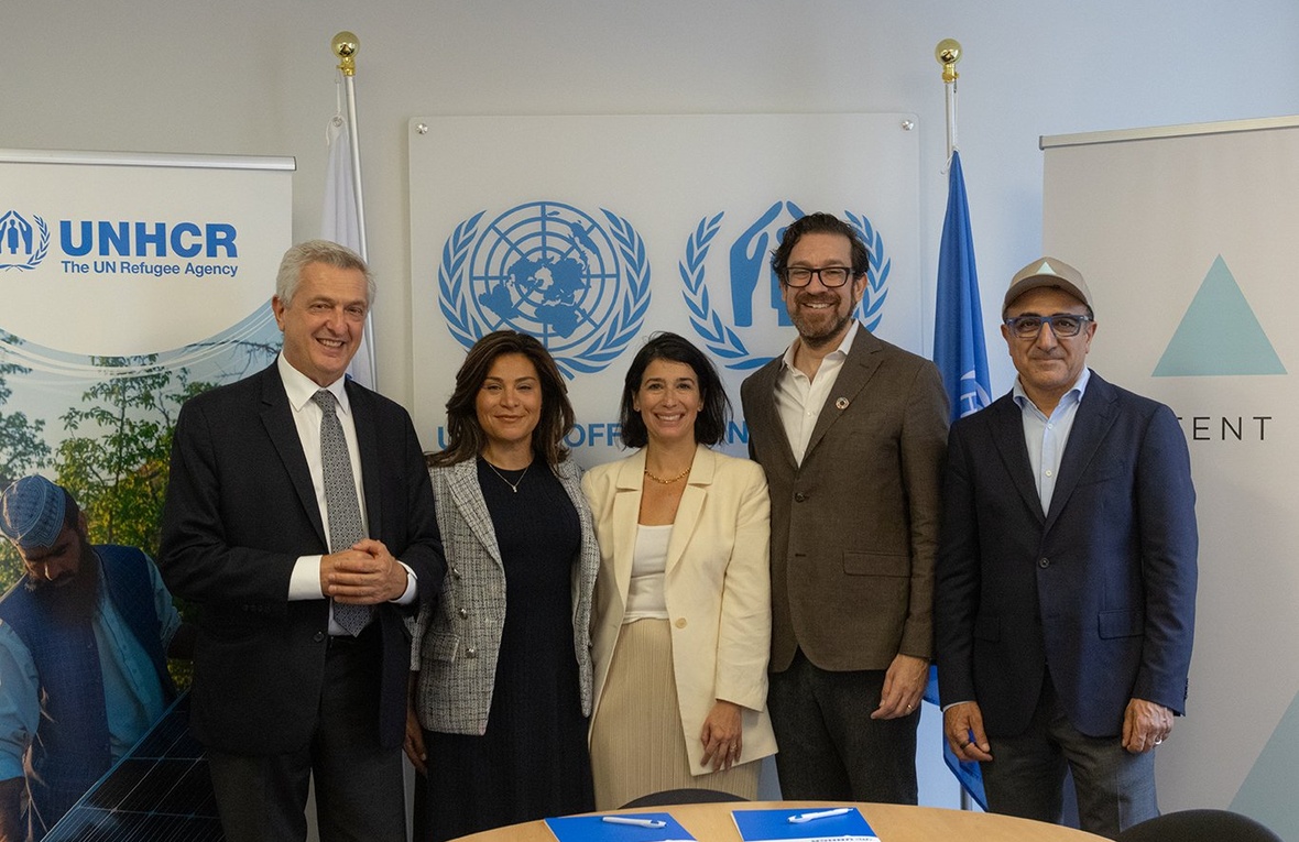 Five people smile and pose for the camera behind a table, with UNHCR banners in the background.