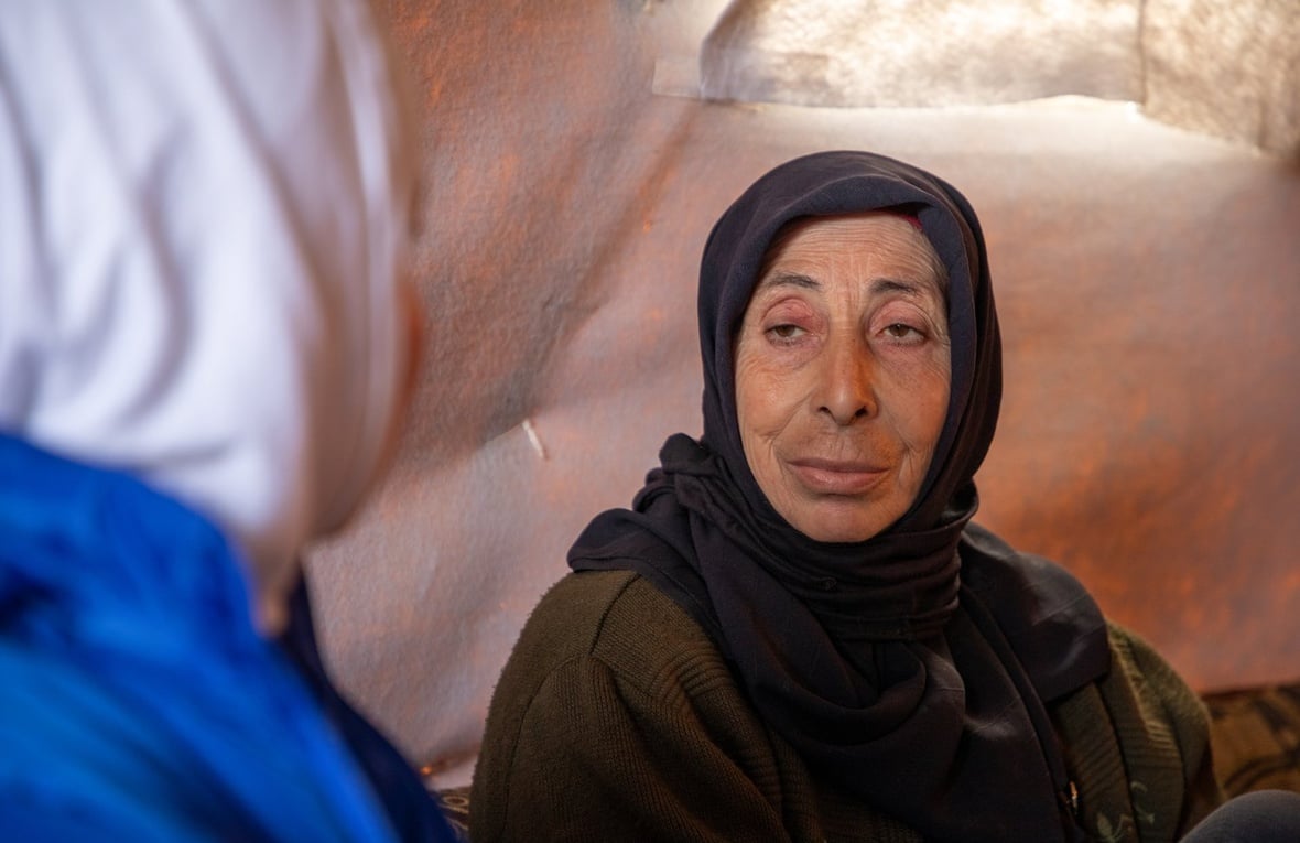 A woman in a dark headscarf sitting inside a tent looks directly at another woman in the foreground wearing a blue jacket and white headscarf whose face is turned away