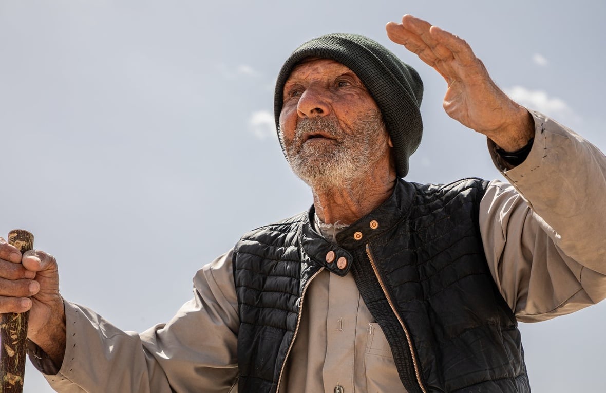 An elderly man wearing a cap and holding a walking stick stands outside beneath a clear sky with his hands raised