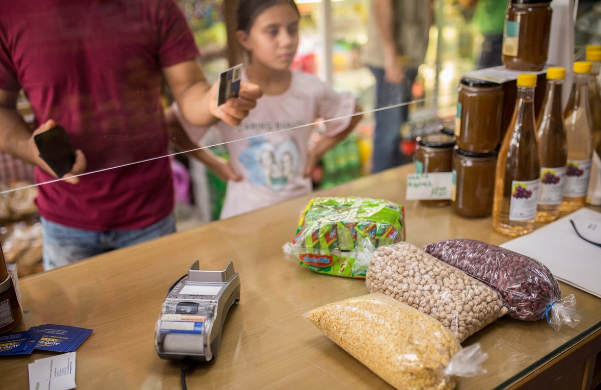 A man stands next to a young girl at the counter of a grocery store holding out a payment card to purchase bags of dried beans and pulses