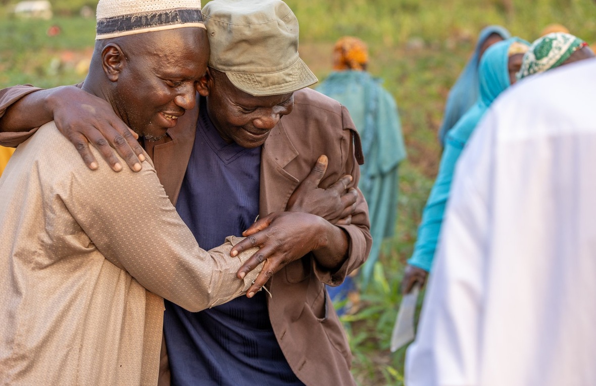 Two men smile and embrace in a field, surrounded by other people