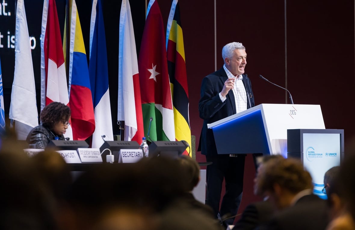A man in a suit gestures while speaking at a lectern in front of a crowd