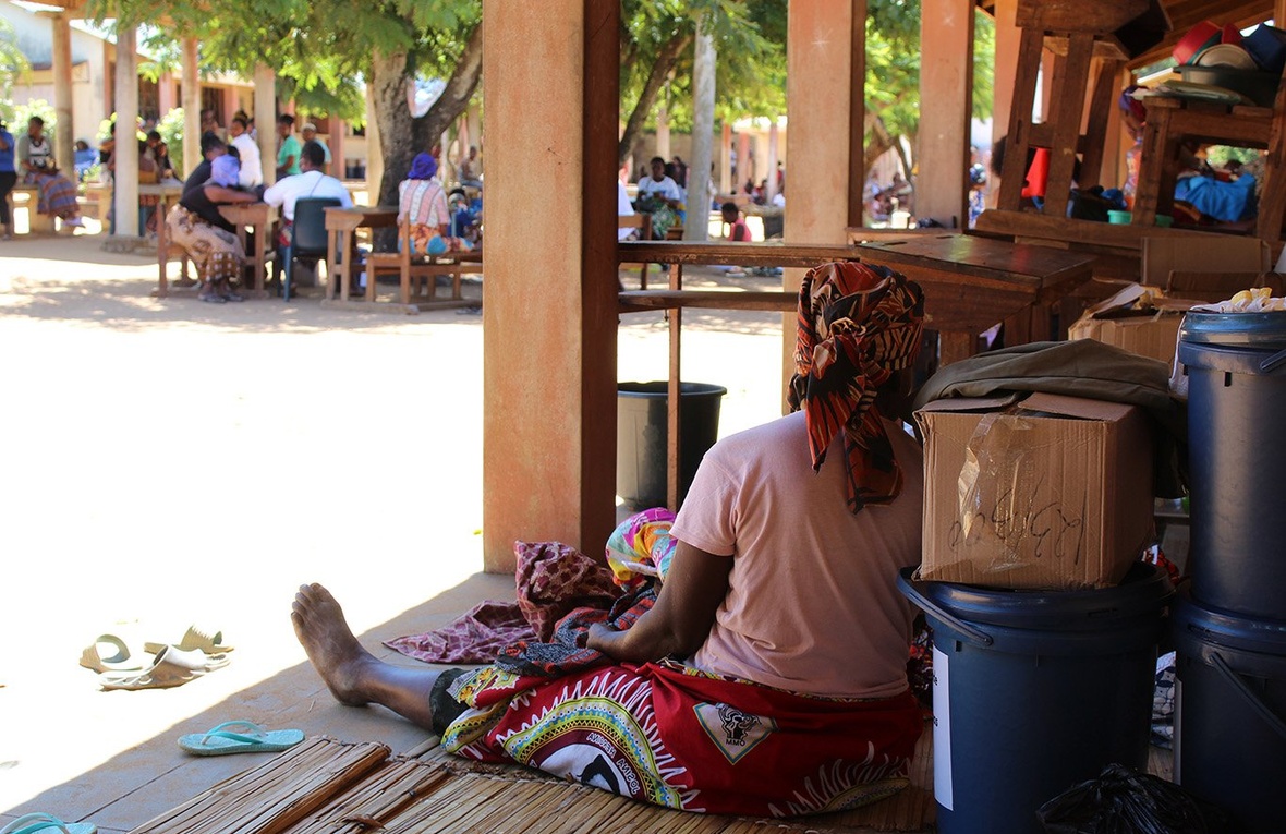 A woman sits in the shade on the wooden terrace of a school, leaning back against boxes of aid supplies 