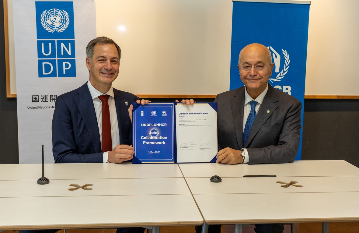 Two smiling men sit at a table, holding up a signed document that reads "UNDP-UNHCR Joint Collaboration Agreement: 2026-2030". Behind them are UNDP and UNHCR banners.