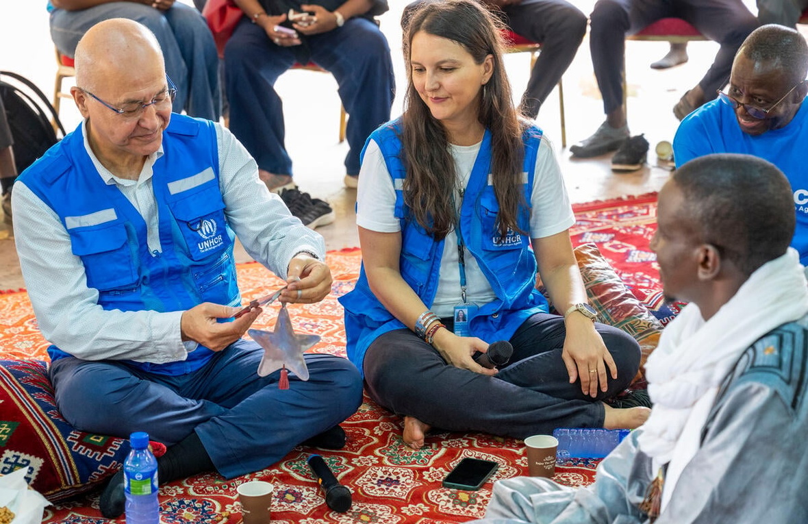 A man and woman wearing blue vests sit cross-legged on a carpet opposite a man 