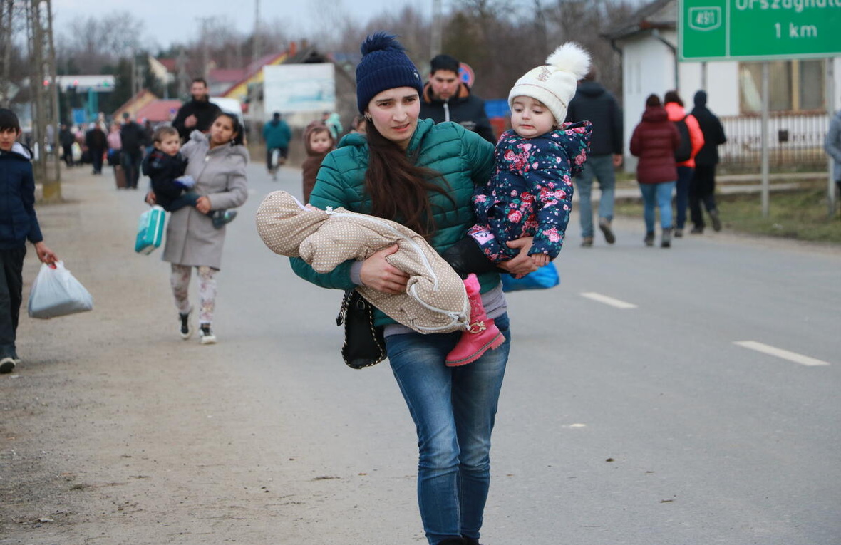 A young woman walks along the side of a road carrying a toddler and a baby in her arms 