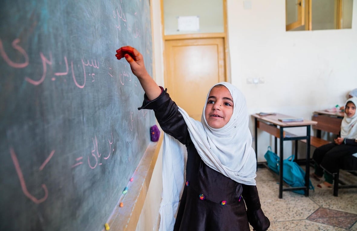 An Afghan girl holds up her arm towards a blackboard in a classroom, as others sit at desks behind her.
