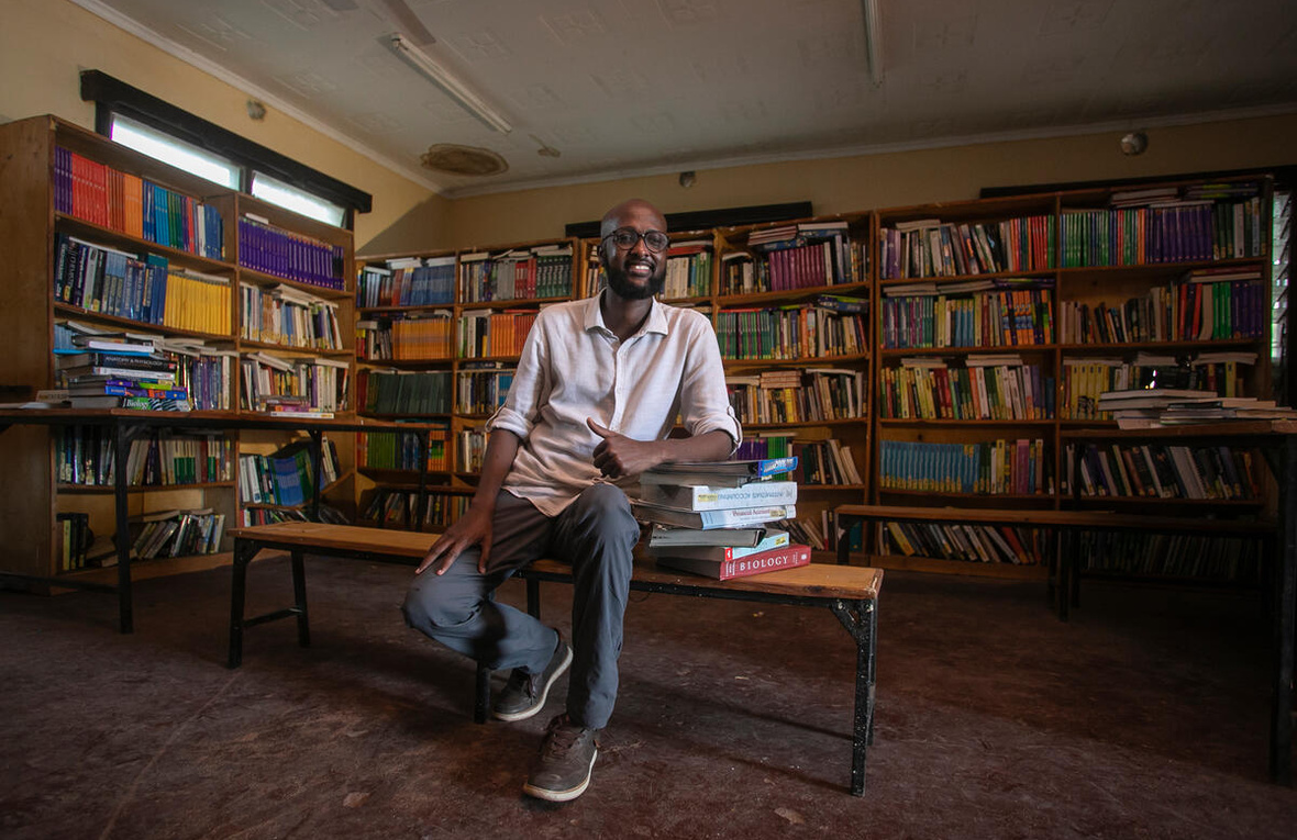 A man sits in a library, surrounded by books and leaning on a pile of books